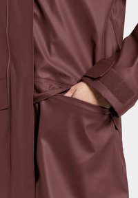 Veste de pluie bordeaux résistante à l'eau avec une texture lisse, fermeture éclair visible et poignet en velcro. Comprend une poche latérale avec une main.