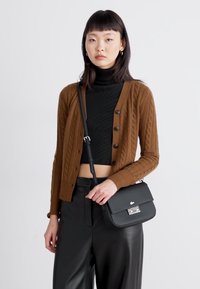 Cardigan en maille torsadée marron avec un col rond, sous un turtleneck noir, et un sac en cuir noir avec des ferrures en argent et un logo, porté avec un pantalon noir.