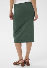 Žena oblečená v tmavozelenej ceruzkovej sukni dĺžky po kolená a hnedých pletených sandáloch na opätku so zadným popruhom, stojaca na jednoduchom pozadí.