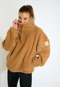 Pullover in pile marrone con colletto alto, ampie tasche e una finitura morbida e texturizzata. Presenta una patch con logo sulla manica sinistra.
