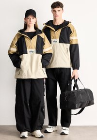 Δύο άτομα φορούν αδιάβροχα μπουφάν με μπλοκ χρωμάτων σε μαύρο, καμηλό και κρεμ, με κορδόνια, μεγάλες τσέπες και έντονο λογότυπο. Φορούν μαύρα cargo παντελόνια και παπούτσια. Το ένα κρατάει μια μαύρη τσάντα duffel.