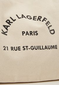 Bolsa de tela en color beige con texto negro en negrita que dice "KARL LAGERFELD," "PARÍS," y "21 RUE ST-GUILLAUME," presentando un diseño sencillo.