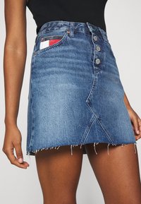 Jeansklänning i medelblått med fransad nederkant. Har hög midja, femknappsstängning och en kontrasterande rutigt fickdetalj.