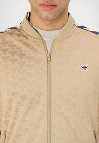 Moški nosi bež vzorčasto jakno z zadrgo, z majhno čebeljim emblemom na levi strani prsnega koša in mornarsko modro črto na rami.