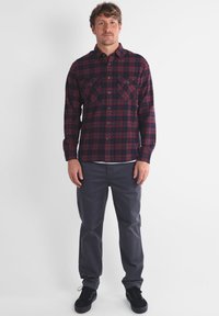 Chemise en flanelle à carreaux de couleur rouge foncé et bleu marine, avec deux poches poitrine, un col à boutons et des manches longues, associée à un pantalon gris.