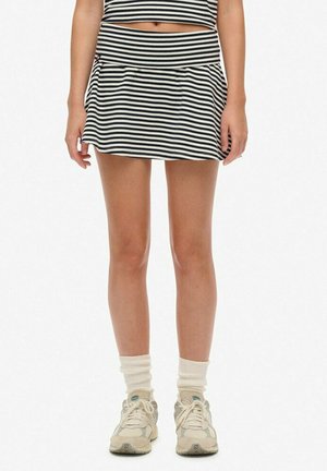 Donna che indossa una gonna e una maglietta a strisce bianche e nere, calze bianche e sneakers beige, in piedi davanti a uno sfondo bianco uniforme.