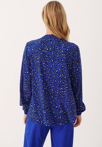 Femme aux cheveux blonds longs portant un chemisier à manches longues imprimé léopard bleu et un pantalon bleu assorti, vue de dos.