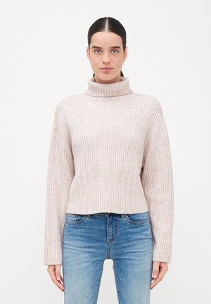 Pull à col roulé côtelé rose clair avec une coupe courte et des épaules tombantes, associé à un jean bleu. Texture douce et coupe décontractée.