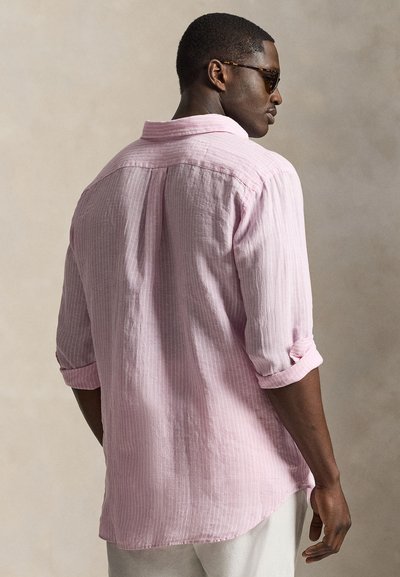 Mees, kes kannab kahvatu roosa triibulist särki, mille varrukad on üles keritud, ja tumedaid päikeseprille, seisab külili neutraalse tausta ees.