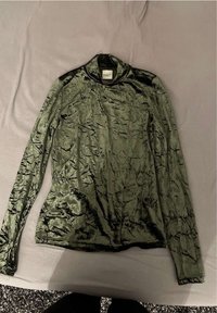 Grünes Langarmoberteil aus glänzendem, strukturiertem Stoff. Verfügt über einen hohen Kragen und komplizierte, gewellte Muster auf der gesamten Oberfläche.