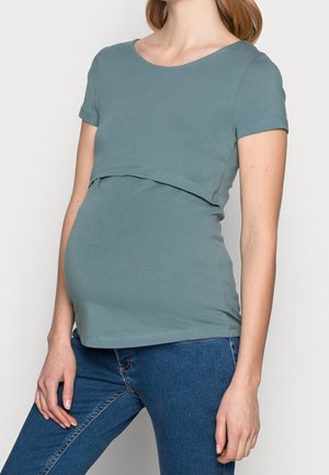 Femme enceinte portant un haut de maternité bleu sarcelle à manches courtes et un jean bleu, la main reposant près de la cuisse.