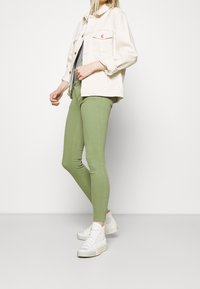 Casual outfit featuring a cream denim jacket, gray top, and green skinny jeans, complemented by white high-top sneakers.