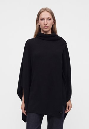 Femme aux longs cheveux blonds portant un poncho noir ample avec un col haut et un pantalon foncé sur un fond blanc uni.