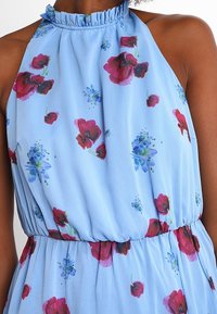 Ärmlös ljusblå klänning med röd och lila blommönster, med hög gathered halsringning och åtsittande midja, bärs av en person med brun hud.