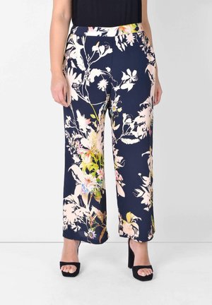 Donna con pantaloni a gamba larga blu navy con stampa floreale rosa e verde e sandali neri con tacco alto e punta aperta.