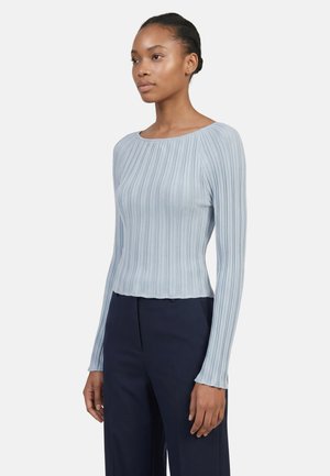 Giovane donna rivolta verso sinistra che indossa una maglia a maniche lunghe azzurro chiaro a coste e pantaloni blu scuro su uno sfondo semplice.
