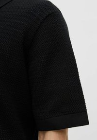Black textured polo shirt featuring short sleeves and a ribbed collar. The fabric has a subtle pattern with a soft, breathable finish.