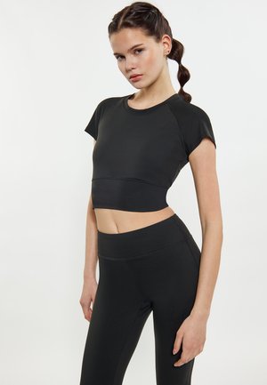Jonge vrouw met gevlochten haar draagt een zwarte cropped sporttop en zwarte leggings met hoge taille tegen een effen witte achtergrond.