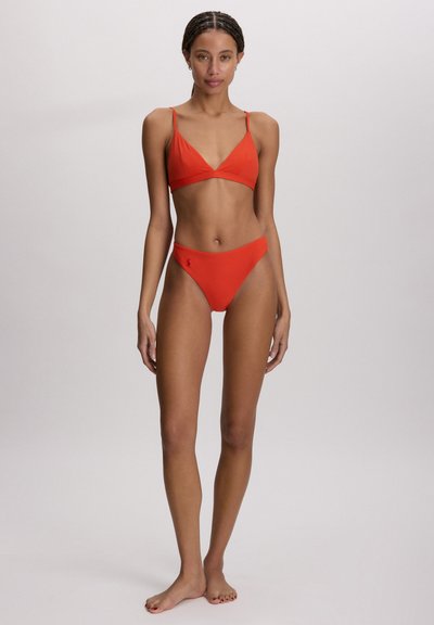 Femeie cu părul împletit, stând desculță, purtând un costum de baie portocaliu din două piese, pe un fundal simplu gri deschis.