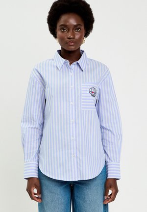 Femme portant une chemise à rayures bleu clair et blanches avec des boutons, brodée d'un coquillage et de "LOVE" sur la poche poitrine, associée à un jean bleu.