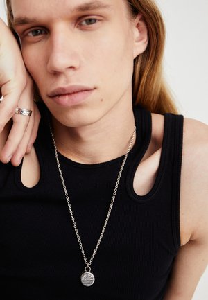 Zwart geribd tanktop, zilveren ketting met ronde hanger met een verhoogd zonontwerp, gedragen met een zilveren ring aan de hand.