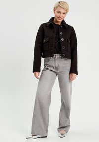 Veste noire cropped avec une finition texturée, grandes poches avant et boutons, associée à un jean taille haute gris clair à jambes larges et des ballerines argentées.