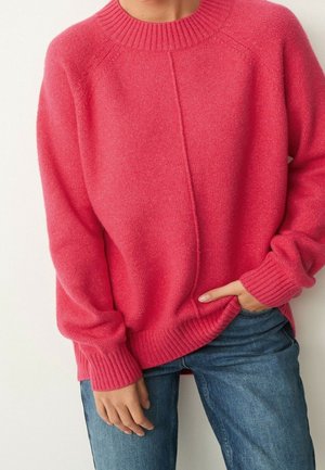 Personne portant un pull à col rond côtelé rose vif à manches longues et un jean bleu, une main dans la poche et l'autre posée de manière détendue le long du corps.