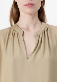 Chemisier beige avec un col en V, des détails froncés aux épaules et un tissu lisse et texturé. Design simple et minimaliste.