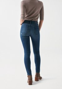Jeans de mezclilla azul de tiro alto con un diseño ajustado, que cuentan con bolsillos traseros y una textura suave, combinados con botas de tacón marrones.