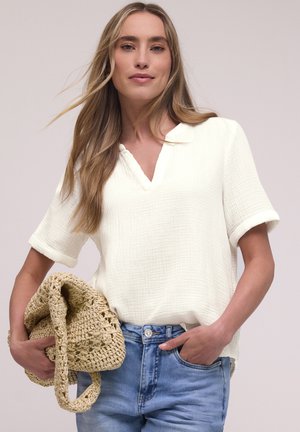 Femme aux longs cheveux blonds portant un chemisier blanc texturé à manches courtes et un jean bleu, tenant un sac tissé beige dans une main.