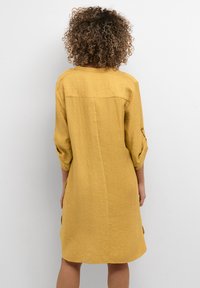 Senapsgul klänning med texturerad tyg, trekvartslånga ärmar, rund halsringning och avslappnad passform, med en bakficka och sidfickor.