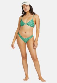 Grön blommig bikini med trekantstop och lågt skurna trosor, tillverkad av mjukt tyg. Modellen har på sig en vit keps och står barfota.