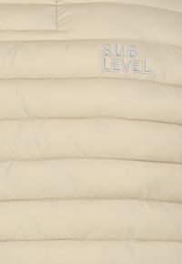 Beige gesteppter Stoff mit horizontaler Naht und den Wörtern "SUB LEVEL" in weißem Faden nahe der oberen rechten Ecke gestickt.