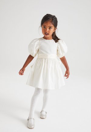 Vestido blanco con mangas cortas abullonadas, cinta en la cintura y falda acampanada. Combinado con medias blancas con estampado de corazones y zapatos plateados brillantes.