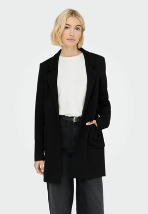 Black textured blazer with a notched collar, open front, and two front pockets. Worn over a white top and paired with black pants.