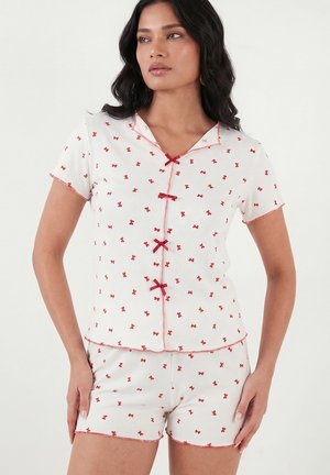 Naine kannab valget lühikese varrukaga pidžaamat, millel on väikesed punased vibu mustrid ja punased vibu aktsendid ees, seisab käed puusal.