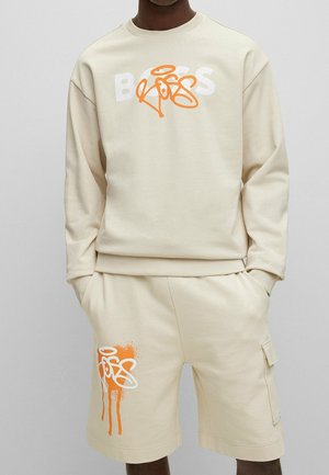 Mand iført beige sweatshirt og matchende shorts med orange og hvidt graffiti-inspireret brand logo, hænderne i lommerne, mod ensfarvet baggrund.