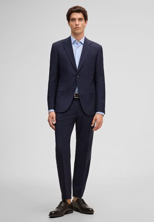 Uomo in piedi con abito blu navy, camicia azzurro chiaro, cintura nera e scarpe eleganti nere davanti a uno sfondo grigio uniforme.