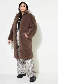 Cappotto in finta pelliccia marrone con colletto alto, design trapuntato e tasche laterali. Abbinato a pantaloni larghi a fantasia e stivali neri chunky.