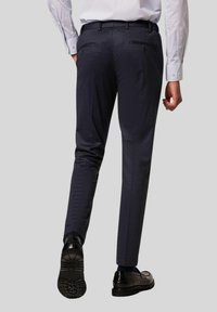 Navy tailored broek met een slim fit, voorzien van twee achterzakken met knoopsluitingen en een subtiele tekstuurstof. Draagt met zwarte schoenen.