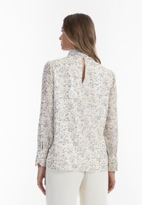 Blomsterbluse i let, gennemsigtigt stof. Har en høj krave, detaljer med nøglehul i ryggen og lange ærmer med manchetter. Farverne inkluderer creme, gul og blå.
