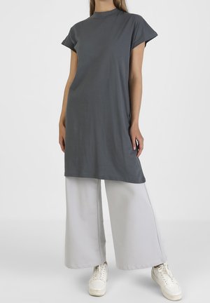 Mujer con túnica de manga corta gris oscuro, pantalones blancos de pierna ancha y zapatillas blancas, de pie contra un fondo claro y liso.