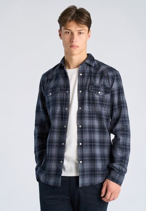 Ung mand står iført en blå og gråternet skjorte med knapper over en hvid t-shirt, foran en ensartet lys baggrund.