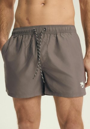 Muž nosí šedé plavky na stažení s bočními kapsami a malým bílým logem na levé noze na jednobarevném pozadí.