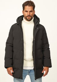 Schwarze Pufferjacke mit Kapuze, Reißverschluss vorne und seitlichen Taschen, über einem weißen Zopfmuster-Pullover getragen, kombiniert mit hellblauen Jeans.