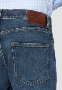 Denim jeans in dark blue with a smooth texture. Features a brown leather waistband patch embossed with "TOMMY HILFIGER" and two back pockets.