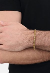 Bransoletka łańcuszkowa w odcieniu złota z przeplatającymi się prostokątnymi ogniwami, noszona na nadgarstku. Bransoletka ma gładkie wykończenie i lekką konstrukcję.