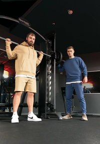 Due uomini in una palestra. Uno indossa un hoodie beige e dei pantaloni corti, l'altro una felpa blu e dei pantaloni da jogging. Attrezzatura da palestra è visibile sullo sfondo.
