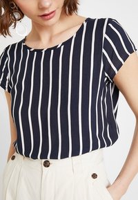 Blusa a righe blu e bianca con scollatura rotonda, maniche corte e tessuto morbido. Abbinata a pantaloni cremosi a vita alta con dettagli a bottoni.