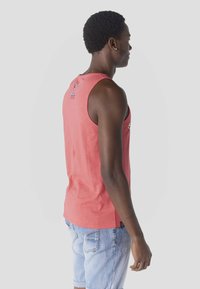 Hombre con cabello corto y rizado, vestido con camiseta sin mangas coral y pantalones cortos de mezclilla azul claro, de perfil contra un fondo liso.